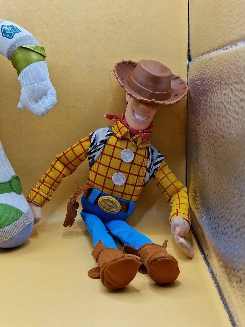 May include: A Woody doll from the Toy Story franchise. The doll is wearing a brown cowboy hat, a yellow plaid shirt, blue pants, and brown boots. A red bandana is around the neck. The doll has a friendly expression.