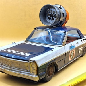 以下が含まれることがあります： 青、白、シルバーのヴィンテージおもちゃのパトカー。サイレンが上に付いています。車にはボンネットと側面に「POLICE」とプリントされ、ドアには番号「77」があります。おもちゃは金属製で、使用感があります。