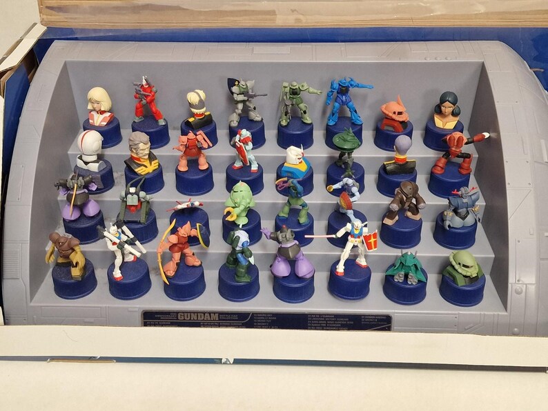 May include: A display case filled with miniature Gundam figures. The figures are on blue bottle caps and are various colors, including red, green, and blue. The display case is gray and has the word "GUNDAM" on it.