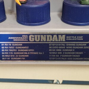May include: A display of Gundam bottle cap figures, featuring a blue base and detailed miniature characters. The image includes a list of Gundam models, celebrating the 25th Anniversary Memorial.