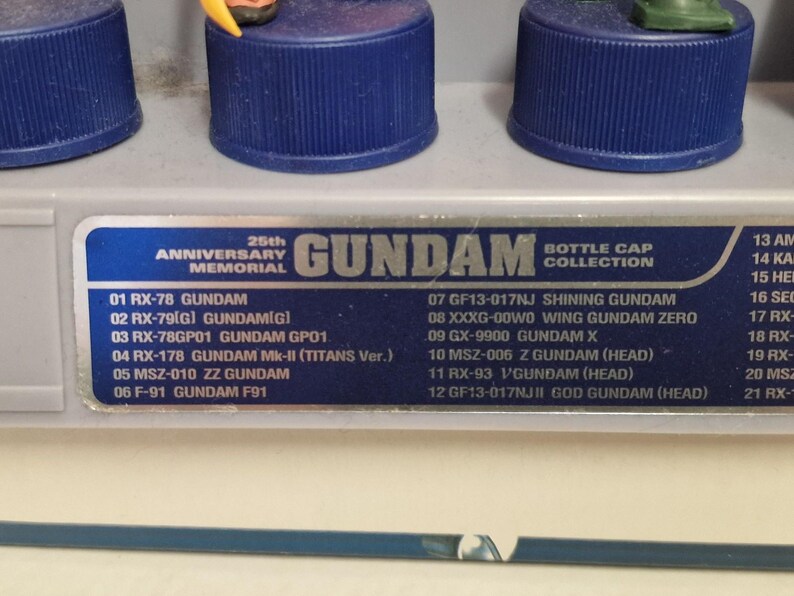 May include: A display of Gundam bottle cap figures, featuring a blue base and detailed miniature characters. The image includes a list of Gundam models, celebrating the 25th Anniversary Memorial.