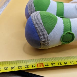May include: Close-up of two white and green plush toy feet with blue and tan accents. The feet have green straps and circular details. A yellow measuring tape with black markings is in the foreground.