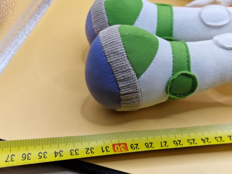 May include: Close-up of two white and green plush toy feet with blue and tan accents. The feet have green straps and circular details. A yellow measuring tape with black markings is in the foreground.