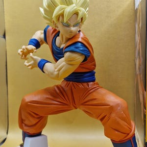 May include: A plastic anime figure of a character with spiky blonde hair, wearing an orange gi with a blue undershirt, blue wristbands, a blue sash, and blue boots with red trim. The figure is in a dynamic combat stance with hands positioned forward.