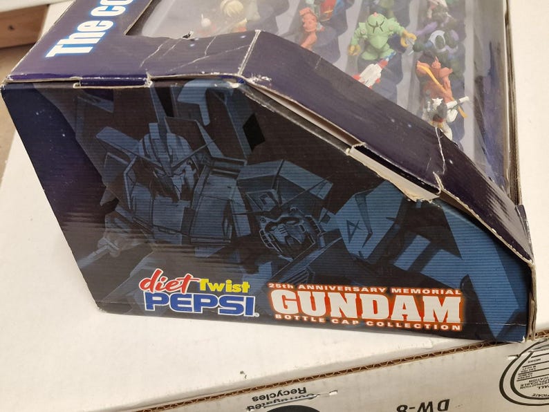 May include: A partially visible box for the 25th Anniversary Memorial Gundam Bottle Cap Collection. The box features a dark blue and black color scheme with images of Gundam robots and the Diet Twist Pepsi logo.