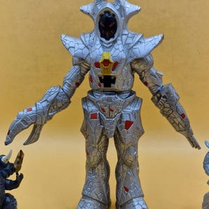 Puede incluir: Figura de acci&oacute;n plateada con apariencia texturizada y agrietada, con detalles en rojo. La figura tiene una cabeza en forma de casco con una cara oscura y est&aacute; posada con los brazos extendidos. Parte de una colecci&oacute;n de figuras de juguete.