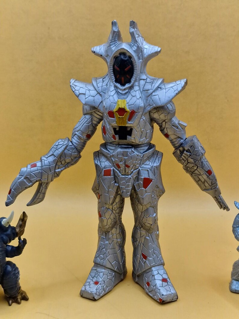 Puede incluir: Figura de acci&oacute;n plateada con apariencia texturizada y agrietada, con detalles en rojo. La figura tiene una cabeza en forma de casco con una cara oscura y est&aacute; posada con los brazos extendidos. Parte de una colecci&oacute;n de figuras de juguete.