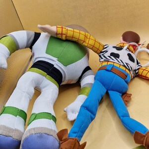 May include: Two plush toy figures, Buzz Lightyear and Woody, from the animated film Toy Story. Buzz is in a white, green, and black spacesuit. Woody wears a cow-print vest, blue pants, and brown boots. A yellow measuring tape is in the foreground.