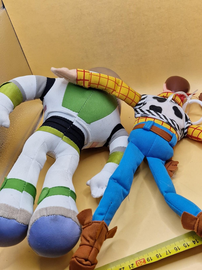 May include: Two plush toy figures, Buzz Lightyear and Woody, from the animated film Toy Story. Buzz is in a white, green, and black spacesuit. Woody wears a cow-print vest, blue pants, and brown boots. A yellow measuring tape is in the foreground.