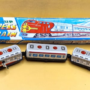 May include: A vintage wind-up toy train set featuring two red and white locomotive engines and a central passenger car. The set includes a rectangular blue and white box labeled "WIND-UP EXPRESS TRAIN" with an illustration of the train.