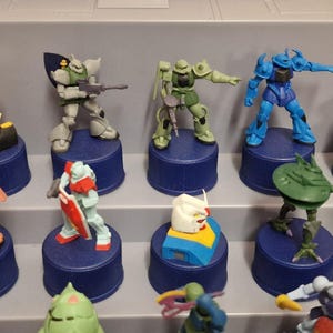 May include: A collection of colorful Gundam model figures displayed on blue bottle caps. The figures are in various poses and colors, including green, blue, red, and white. The figures are arranged on a gray shelf.