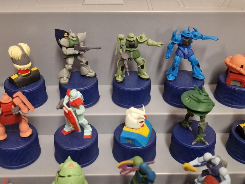 May include: A collection of colorful Gundam model figures displayed on blue bottle caps. The figures are in various poses and colors, including green, blue, red, and white. The figures are arranged on a gray shelf.