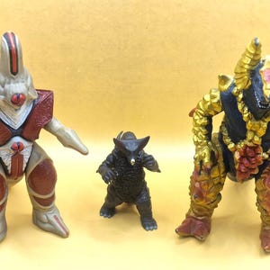 May include: Three standing monster figurines. The left figure is beige with dark red armor plates. The center figure is small, black, and textured. The right figure is black with gold, textured, rock-like armor and a prominent golden horn structure.