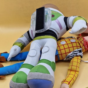 May include: Two plush toy figures, Buzz Lightyear and Woody, from the Toy Story franchise. Buzz Lightyear is wearing a white, green, and purple spacesuit. Woody is wearing a yellow plaid shirt and blue jeans.