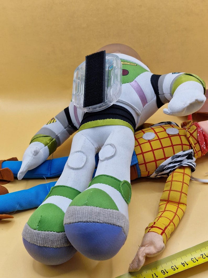 May include: Two plush toy figures, Buzz Lightyear and Woody, from the Toy Story franchise. Buzz Lightyear is wearing a white, green, and purple spacesuit. Woody is wearing a yellow plaid shirt and blue jeans.
