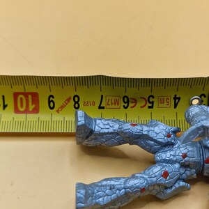 Puede incluir: Una figura de acci&oacute;n gris con detalles rojos, de aproximadamente 9 cm de largo, se coloca junto a una cinta m&eacute;trica amarilla. La figura tiene una apariencia texturizada, similar a la piedra. La cinta muestra medidas m&eacute;tricas.