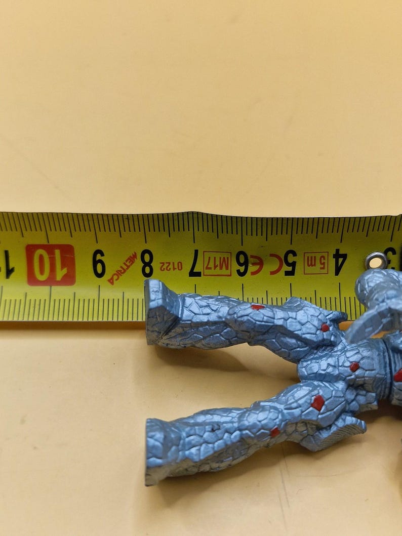 Puede incluir: Una figura de acci&oacute;n gris con detalles rojos, de aproximadamente 9 cm de largo, se coloca junto a una cinta m&eacute;trica amarilla. La figura tiene una apariencia texturizada, similar a la piedra. La cinta muestra medidas m&eacute;tricas.