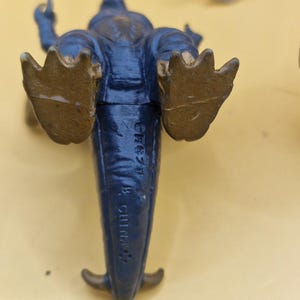 Puede incluir: Una figura de juguete de dinosaurio azul con pies y cola dorados. Se ve la parte inferior del juguete, con el texto "CH87F B CHINA" impreso. La figura est&aacute; sobre una superficie amarilla.