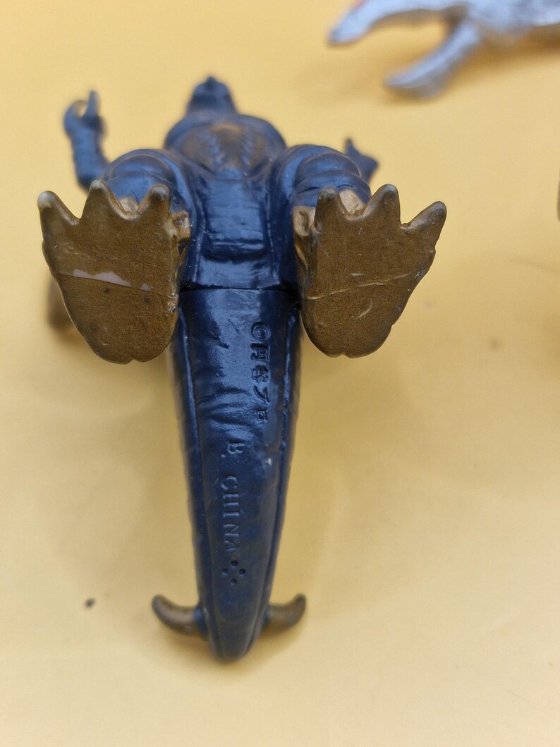 Puede incluir: Una figura de juguete de dinosaurio azul con pies y cola dorados. Se ve la parte inferior del juguete, con el texto "CH87F B CHINA" impreso. La figura est&aacute; sobre una superficie amarilla.
