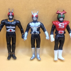 Peut inclure: Cinq figurines de personnages costumés, chacune avec une palette de couleurs unique. Les figurines sont principalement noires avec des accents argentés, rouges et bleus. Elles ont des casques et des ceintures, et sont alignées.