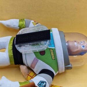 May include: A Buzz Lightyear plush toy, featuring a white and green spacesuit with black accents. The toy has a clear helmet and the word "LIGHTYEAR" is visible on the arm. The toy is lying on a yellow surface.