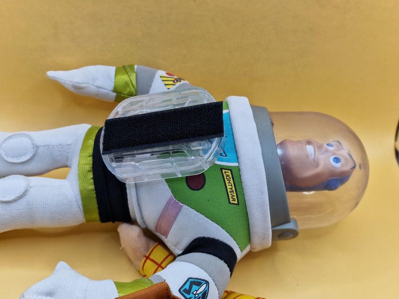 May include: A Buzz Lightyear plush toy, featuring a white and green spacesuit with black accents. The toy has a clear helmet and the word "LIGHTYEAR" is visible on the arm. The toy is lying on a yellow surface.