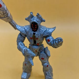 Puede incluir: Figura de acci&oacute;n plateada de una criatura humanoide con apariencia rocosa texturizada. La figura tiene detalles rojos y sostiene un arma. La figura est&aacute; posada sobre un fondo amarillo.