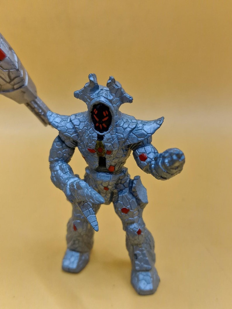 Puede incluir: Figura de acci&oacute;n plateada de una criatura humanoide con apariencia rocosa texturizada. La figura tiene detalles rojos y sostiene un arma. La figura est&aacute; posada sobre un fondo amarillo.