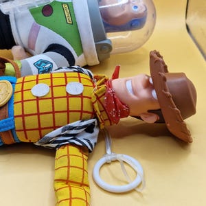 May include: A close-up of two Toy Story character dolls. Woody, the cowboy, is in the foreground, wearing a yellow plaid shirt, blue pants, and a brown hat. Buzz Lightyear is in the background, wearing a white and green space suit.