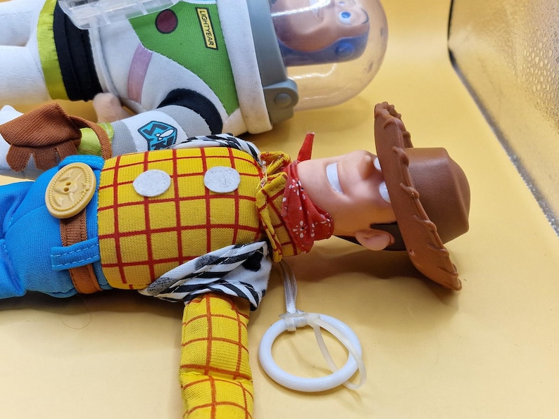 May include: A close-up of two Toy Story character dolls. Woody, the cowboy, is in the foreground, wearing a yellow plaid shirt, blue pants, and a brown hat. Buzz Lightyear is in the background, wearing a white and green space suit.