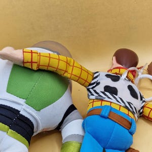 May include: Two plush toy figures, Woody and Buzz Lightyear, from the animated film Toy Story. Woody is wearing a yellow plaid shirt, blue jeans, and a cow-print vest. Buzz Lightyear is in a white and green spacesuit. A brown cowboy hat is also visible.