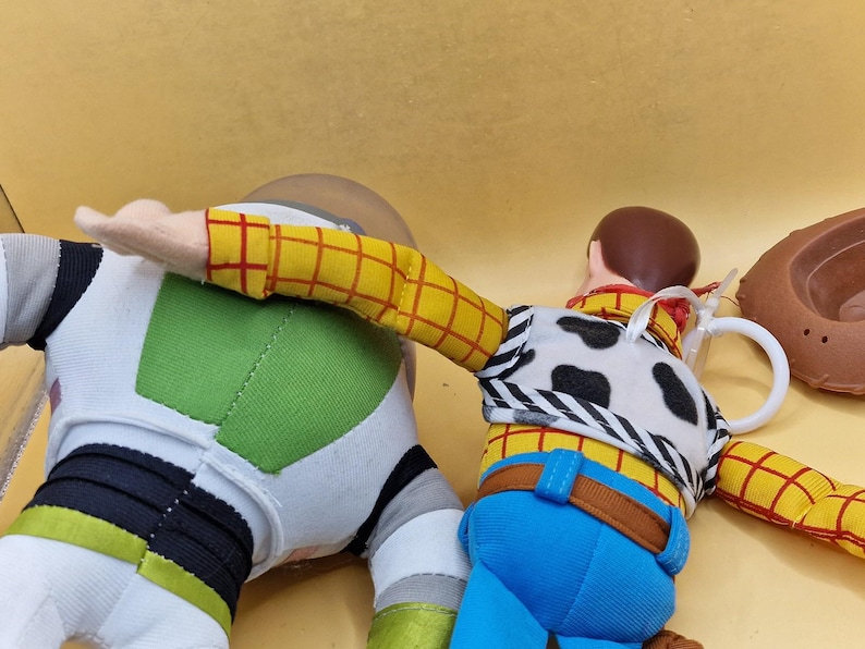 May include: Two plush toy figures, Woody and Buzz Lightyear, from the animated film Toy Story. Woody is wearing a yellow plaid shirt, blue jeans, and a cow-print vest. Buzz Lightyear is in a white and green spacesuit. A brown cowboy hat is also visible.
