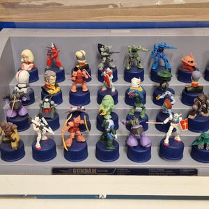 May include: A collection of Gundam character figurines displayed in a tiered, gray plastic case. The figurines are colorful, with various poses and designs, each mounted on a blue base. The word "GUNDAM" is printed on the case.