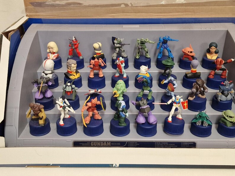 May include: A collection of Gundam character figurines displayed in a tiered, gray plastic case. The figurines are colorful, with various poses and designs, each mounted on a blue base. The word "GUNDAM" is printed on the case.