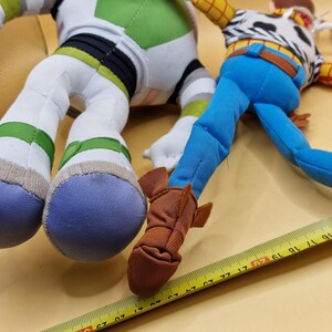 May include: Close-up of two plush toy figures: Buzz Lightyear and Woody. Buzz Lightyear has white, green, and black accents. Woody is wearing a cowboy outfit with blue pants and brown boots. A yellow measuring tape is in the foreground.