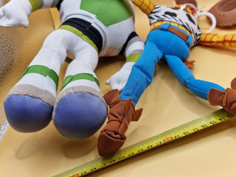 May include: Close-up of two plush toy figures: Buzz Lightyear and Woody. Buzz Lightyear has white, green, and black accents. Woody is wearing a cowboy outfit with blue pants and brown boots. A yellow measuring tape is in the foreground.
