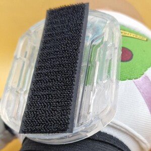 May include: A clear plastic accessory with a black hook-and-loop fastener. The accessory is attached to a white and green fabric with the word "LIGHTYEAR" printed on it. The background is a soft yellow.