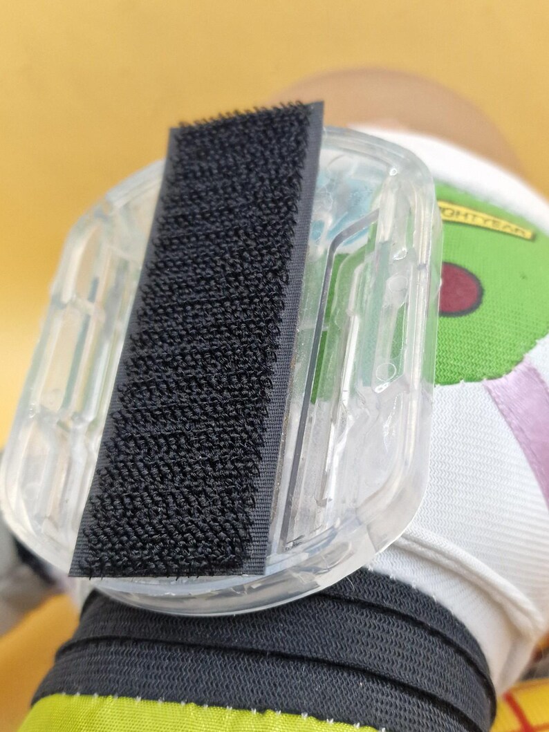 May include: A clear plastic accessory with a black hook-and-loop fastener. The accessory is attached to a white and green fabric with the word "LIGHTYEAR" printed on it. The background is a soft yellow.