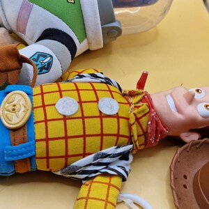 May include: A close-up of a Woody doll from the Toy Story movie, with a yellow plaid shirt, blue pants, and a red bandana. A brown cowboy hat is next to the doll. Part of a Buzz Lightyear doll is also visible.