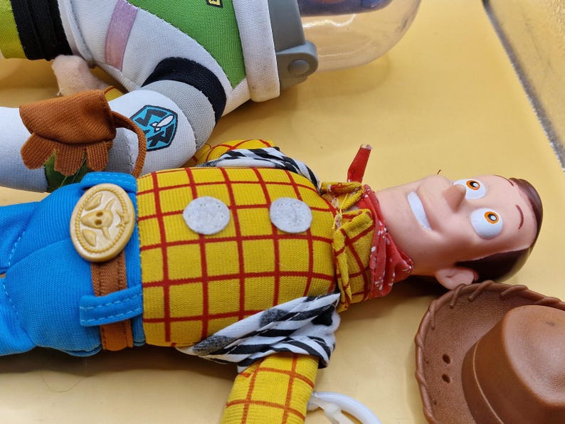 May include: A close-up of a Woody doll from the Toy Story movie, with a yellow plaid shirt, blue pants, and a red bandana. A brown cowboy hat is next to the doll. Part of a Buzz Lightyear doll is also visible.