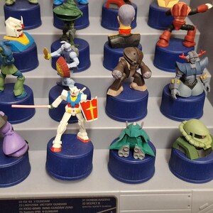 May include: A collection of colorful Gundam figures displayed on blue bottle caps. The figures are in various poses and designs, with some holding weapons or shields. The display is on a tiered shelf.