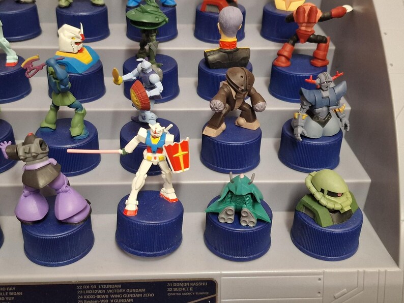 May include: A collection of colorful Gundam figures displayed on blue bottle caps. The figures are in various poses and designs, with some holding weapons or shields. The display is on a tiered shelf.