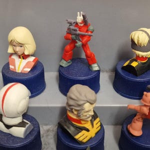 May include: A collection of small, colorful figurines, including busts and action figures, displayed on blue bottle caps. The figurines feature various characters with detailed designs and vibrant colors, such as red, white, and yellow.