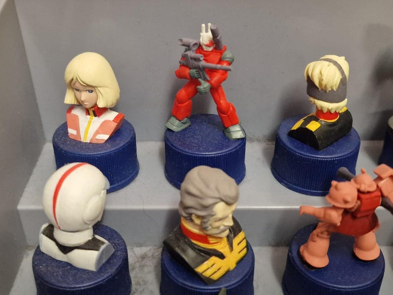 May include: A collection of small, colorful figurines, including busts and action figures, displayed on blue bottle caps. The figurines feature various characters with detailed designs and vibrant colors, such as red, white, and yellow.