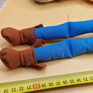 May include: Close-up of a toy doll's legs. The doll's legs are blue, with brown boots. A yellow ruler with black markings is in the foreground, measuring in inches.