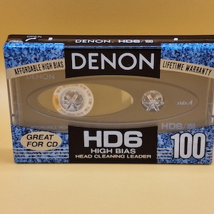 Op de afbeelding: Een blauwe en zilveren cassetteband met de tekst "DENON HD6 100 High Bias", "Great for CD" en "Head Cleaning Leader".