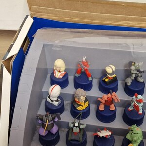 May include: A collection of small, colorful figurines displayed in a clear plastic case. The figurines, featuring various characters, are mounted on dark blue bases. The case is partially open, revealing a cardboard box with blue accents.