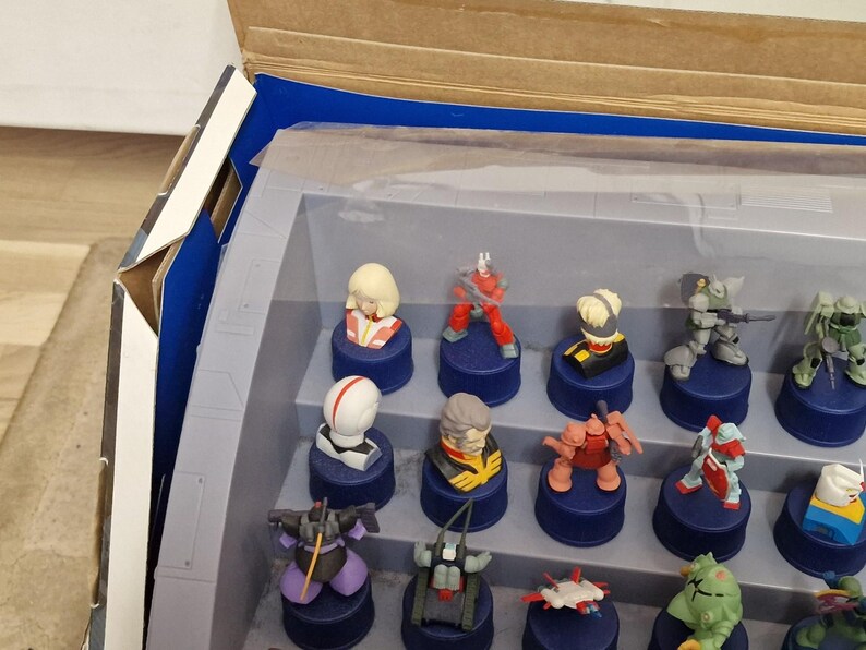 May include: A collection of small, colorful figurines displayed in a clear plastic case. The figurines, featuring various characters, are mounted on dark blue bases. The case is partially open, revealing a cardboard box with blue accents.