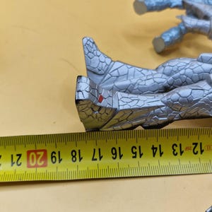 Puede incluir: Una figura de color plateado con una apariencia texturizada similar a la piedra. Una cinta m&eacute;trica amarilla se coloca junto a la figura, mostrando medidas de 12 a 21 cm. La figura parece ser parte de una figura m&aacute;s grande.