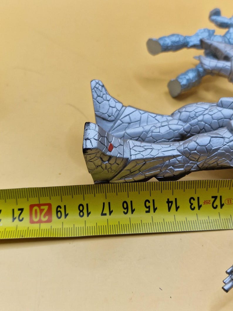 Puede incluir: Una figura de color plateado con una apariencia texturizada similar a la piedra. Una cinta m&eacute;trica amarilla se coloca junto a la figura, mostrando medidas de 12 a 21 cm. La figura parece ser parte de una figura m&aacute;s grande.